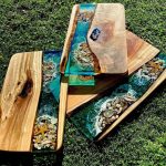 Handcrafted wooden boards with ocean-themed designs created using clear epoxy resin. The clear epoxy embeds seashells and starfish, giving the boards a vibrant and intricate appearance, displayed on green grass.